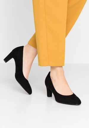 Czarne zamszowe szpilki z okrągłym noskiem, średniej wysokości blokowy obcas i gładka tekstura, zestawione z żółtymi spodniami.