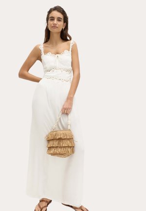 Kobieta w białej, bezrękawnikowej sukience maxi z koronkowymi detalami, trzymająca beżową torebkę z frędzlami, nosząca sandały i minimalistyczną biżuterię.