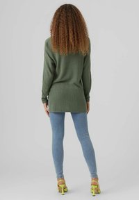 Žena s kudrnatými vlasy, oblečená v dlouhém olivově zeleném svetru a světlých modrých džínách, stojí zády k fotoaparátu a předvádí žluté podpatky.