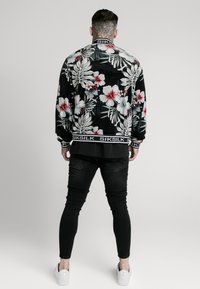 Květovaná bomber bunda z černé látky s bílými tropickými vzory, červenými a růžovými květy a černým žebrovaným límcem a lemem s logem.