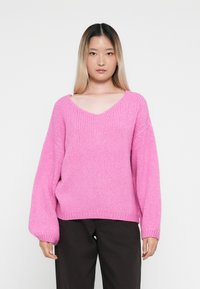 Maglione rosa lavorato a maglia con scollatura a V, vestibilità oversize e maniche a palloncino. Texture spessa e orlo a coste. Abbinato a pantaloni scuri.