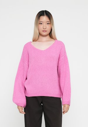 Roze gebreide trui met een V-hals, oversized pasvorm en ballonmouwen. Dikke textuur en geribbelde zoom. Gekaapt met donkere broeken.