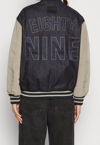 Bomberjacka med kropp i mörk denim, beige ärmar och stor "NITTA NIO" broderad på baksidan. Har randiga ribbade muddar och nederkant.