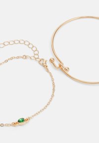 Set di bracciali in tonalità oro, composto da un bangle liscio e una catena con un accento di pietra verde, che mette in evidenza texture e forme diverse.