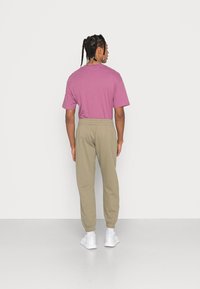 Purpura īsām piedurknēm T-krekls un gaiši khaki sporta bikses ar elastīgām kājām, valkāts ar baltām sporta kurpēm. Vienkārša fona.