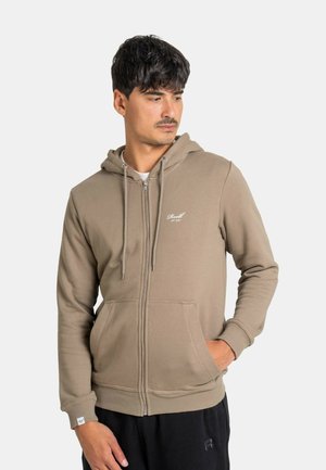 Mann trägt einen beigen Hoodie mit Reißverschluss und Fronttaschen, schaut nach links, eine Hand in der Tasche, kurze dunkle Haare.