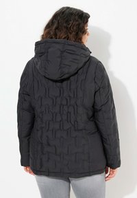 Svart pufferjacka med huva, stoppad textur, elastiska ärmslut och en något rundad nederkant. Synlig sömdekoration på ryggen.