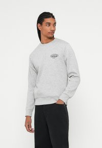 Szürke crewneck pulóver puha, texturált anyagból készült, bal mellen egy kis fekete logóval és bordázott ujjakra és aljára. Fekete nadrággal párosítva.