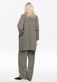 Persona rubia que lleva un blazer de cuadros extragrande y pantalones anchos a juego, de pie de espaldas, sobre un fondo blanco liso.