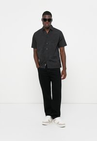 Short-sleeve black shirt with a subtle pattern, button front, paired with black pants and white sneakers. Model wears dark sunglasses.