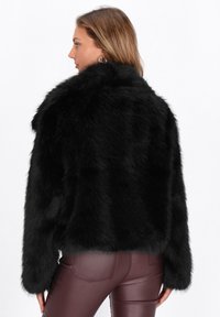 Kvinde set sideværts iført en sort faux fur jakke og mørkeblå læderbukser med hoop øreringe mod en neutral baggrund.