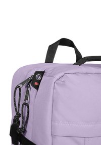 Lila tygryggsäck med en strukturerad yta, med svarta remmar, en dragkedja, och en dekorativ röd accent nära dragkedjans drag.