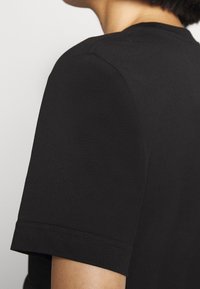 Gros plan d'une personne portant un t-shirt noir uni à manches courtes, montrant l'épaule et le haut du bras.