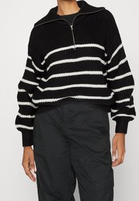 Gestreifter, schwarz-weißer Strickpullover mit hohem Kragen und halbem Reißverschluss vorne; verfügt über lockere, lange Ärmel und einen cropped Saum.