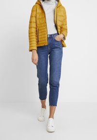 Chaqueta acolchada amarilla con capucha, jersey de cuello vuelto gris, jeans ajustados azules y zapatillas blancas. La chaqueta presenta costuras horizontales y bolsillos con cremallera.