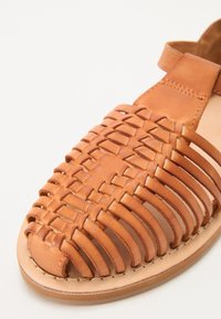 Brune lær sandal med flettet design, åpen tå, flat såle og ankelrem. Glatt tekstur med synlige sømdetaljer.