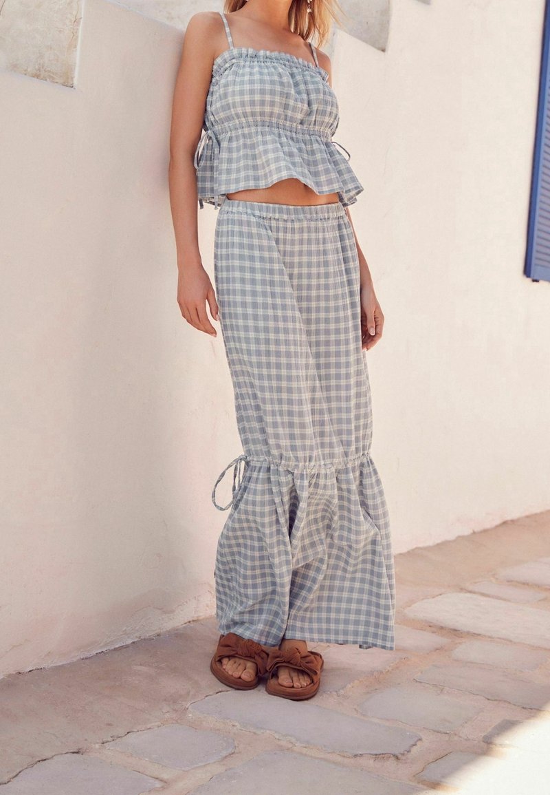 Femme portant une tenue deux pièces à carreaux bleu clair et blanc avec un crop top et un pantalon large, debout sur un pavé en pierre près d’un mur blanc.