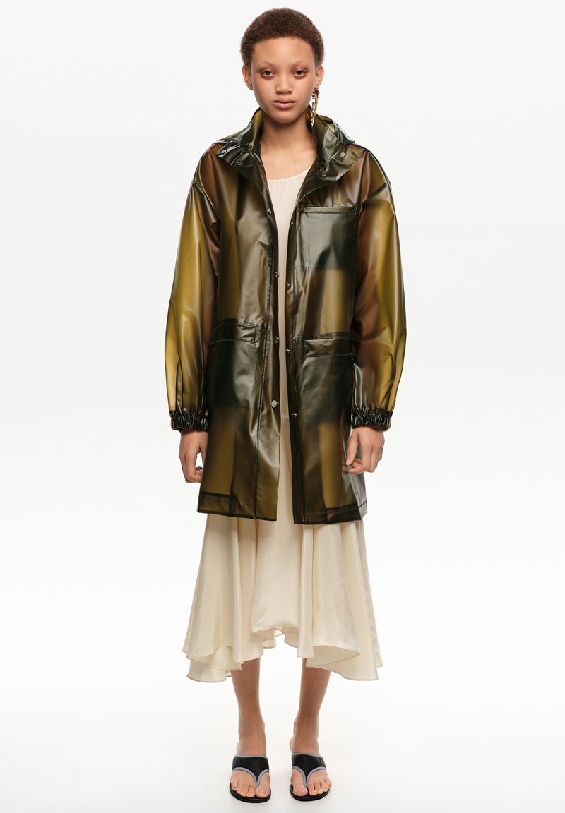 Jeune personne portant un imperméable translucide olive foncé sur une robe crème à ourlet asymétrique et des sandales noires à anneau d'orteil, debout devant un fond blanc.