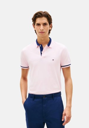 Joven con una camiseta polo rosa claro con cuello y puños azul marino, combinada con pantalones azul oscuro, de pie con una mano en el bolsillo.