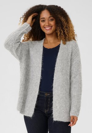 Une femme souriante touche ses cheveux bouclés en portant un cardigan gris clair ouvert sur le devant, par-dessus un haut bleu marine et un jean foncé.