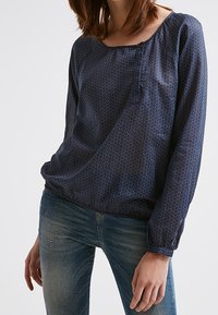 Námořnická modrá halenka s dlouhým rukávem s kulatým výstřihem, detaily s knoflíky a bílými maličkými puntíky na lehkém, mírně průhledném materiálu.