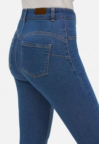 Jeans de mezclilla azul de tiro alto con un diseño ajustado, costuras clásicas, dos bolsillos traseros y una etiqueta en la cintura.