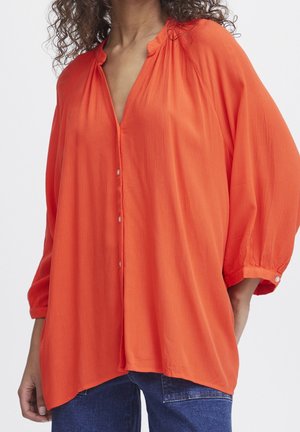 Kvinde iført løs, knap-op, lys orange bluse med trekvart lange pufærmer og blå jeans, stående mod en ensfarvet baggrund.