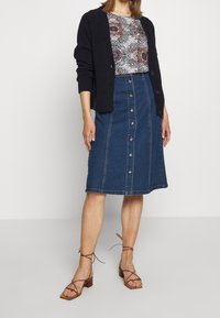 Falda midi de mezclilla con cierre de botón en la parte delantera, que presenta una cintura alta, costura central y una leve silueta en A. Combinada con una camisa estampada y un cárdigan.
