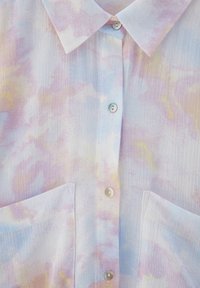 Chemise boutonnée en tissu léger, présentant un motif aquarelle pastel en rose, bleu et jaune. Deux poches avant et boutons perlés.