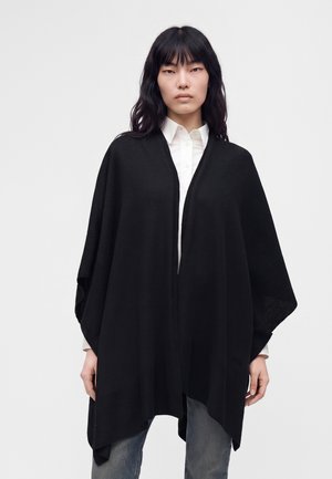 Femme portant un poncho noir ouvert sur une chemise blanche à boutons et un jean bleu, debout devant un fond clair uni.
