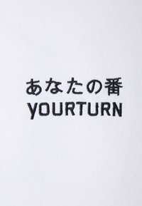 Caractères japonais brodés en noir et texte "YOURTURN" sur tissu blanc, centrés et clairement visibles.