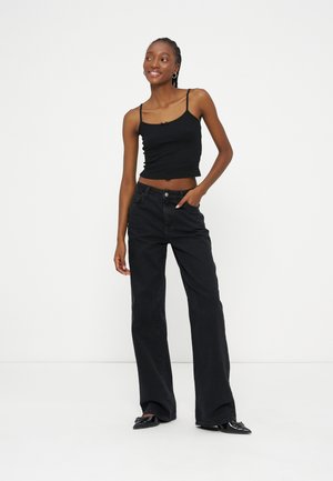 Jeune femme souriante, portant un top court noir, un jean noir à jambes larges et des chaussures noires à bouts pointus, debout devant un fond blanc.