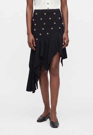 Femme portant une jupe asymétrique noire avec des œillets dorés et un ourlet à volants, associée à des ballerines noires à bout carré.