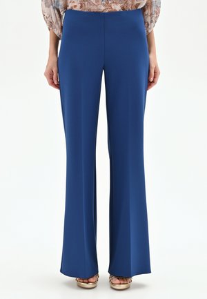 Pantalon bleu à jambes droites porté avec un chemisier beige à motifs et des sandales compensées à bout ouvert dorées sur fond blanc.