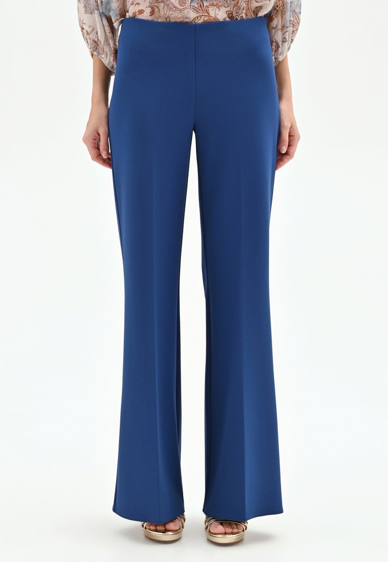 Pantalon bleu à jambes droites porté avec un chemisier beige à motifs et des sandales compensées à bout ouvert dorées sur fond blanc.