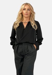 Czarna bluzka z drapowanym dekoltem i długimi rękawami; złoty łańcuszek akcentujący przy kołnierzu. Szare spodnie w prążki z wysokim stanem i zakładkami.