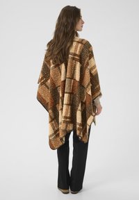 Poncho à carreaux dans des nuances de marron, crème et vert foncé, fabriqué à partir d'un matériau texturé et doux, avec une coupe ample et des bords arrondis.