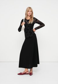 Schwarzes Langarmoberteil mit floralem Muster, kombiniert mit einem schwarzen, gefältelten Rock. Das Modell trägt bordeauxfarbene Ballerinas und hält eine weiße Handtasche.