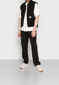 Svart ärmlös väst över en vit t-shirt, tillsammans med svarta cargobyxor och ljusgrå sneakers. Vesten har en dragkedja framtill och en logopatch.