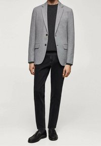 Blazer grigio chiaro con bottoni marroni, abbinato a un maglione scuro e jeans neri. L'outfit presenta eleganti mocassini neri per un look coerente.