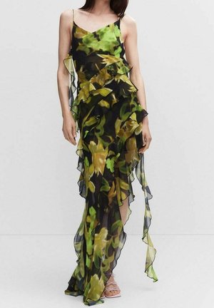 Femme portant une robe noire transparente avec des motifs floraux verts et jaunes et des volants superposés, posant devant un fond blanc uni.