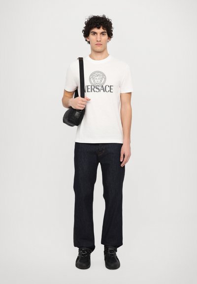 Hvid bomulds t-shirt med et gråt Versace-logo, mørkeblå denimjeans, sort bæltetaske og sorte sneakers med teksturerede detaljer.