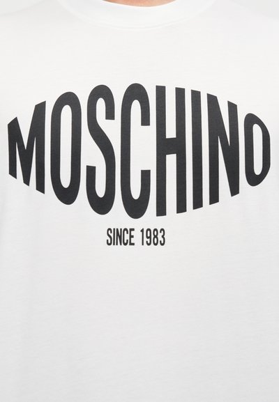 Λευκό βαμβακερό T-shirt με έντονη μαύρη τυπογραφία: "MOSCHINO" κυρτωμένο πάνω από το "SINCE 1983." Ομ αλή υφή και σχεδιασμός με στρογγυλή λαιμόκοψη.