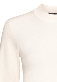 Detailní záběr na pletený svetr krémové barvy s žebrovaným stojáčkem a dlouhými rukávy.