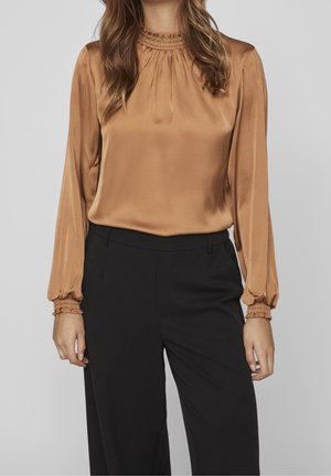 Woman wearing a long-sleeve, camel-colored satin blouse with smocked neckline and cuffs, paired with high-waisted black trousers.