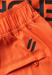 Orangefarbene Sportshorts aus strukturiertem Stoff, mit elastischem Bund und einem schwarzen Logoakzent. Verfügt über eine sichere Seitentasche mit einem orangefarbenen Toggle.
