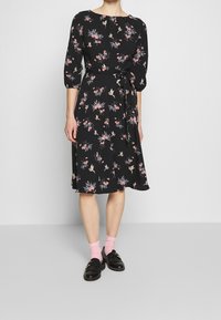 Svart klänning med blommönster, trekvartsärmar och knytning i midjan. Kjol med utsvängd form och längd strax ovanför knät. Kompletterad med rosa strumpor.