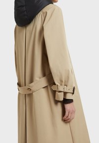 Beige trenchcoat med en svart, puffig huva. Har bälte i midjan, manschettremmar och en jämn textur. Inga synliga knäppningar visas.