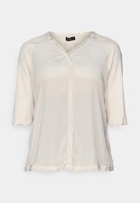 Blouse beige clair à manches courtes avec décolleté en V, texture douce et coupe légèrement ample. Présente une patte de boutonnage centrale et des finitions brutes.
