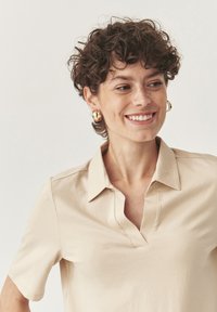 Camisa polo beige con escote en V y mangas cortas, hecha de una tela suave. Los detalles destacados incluyen acentos cosidos y un corte relajado.